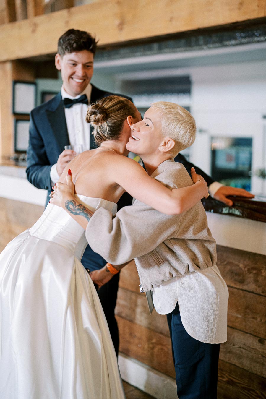 A bride in a white wedding dress embraces a woman with short blonde hair, while a man in a tuxedo smiles in the background at a rustic wedding venue.