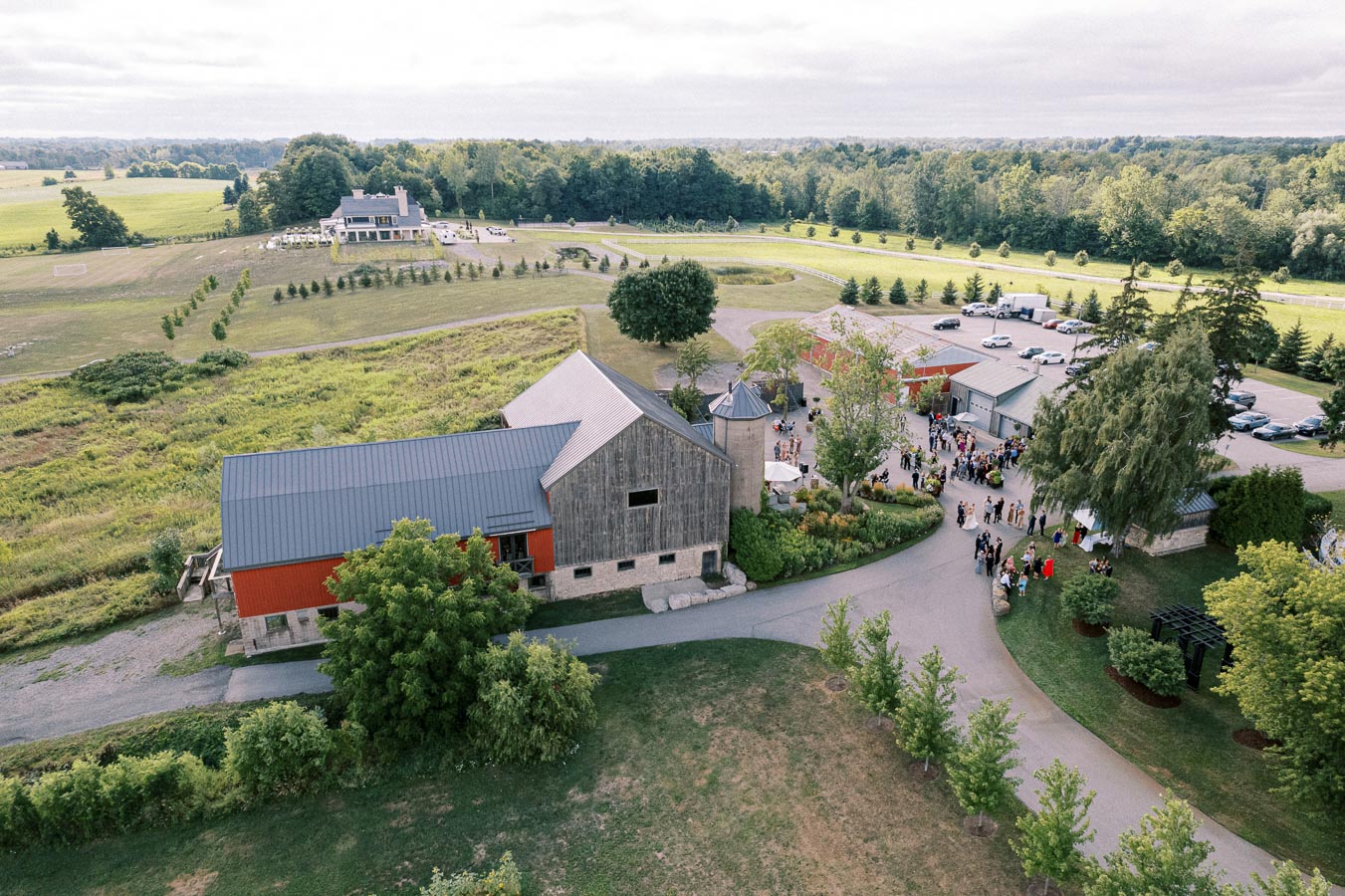 Aerial view of a scenic countryside wedding venue featuring rustic barns surrounded by lush greenery and open fields. Gathering of guests visible in the courtyard, with ample parking and an inviting landscape, perfect for outdoor events.