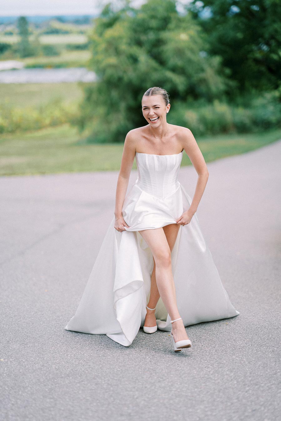 Bride in a strapless white wedding dress laughing on a scenic outdoor path.