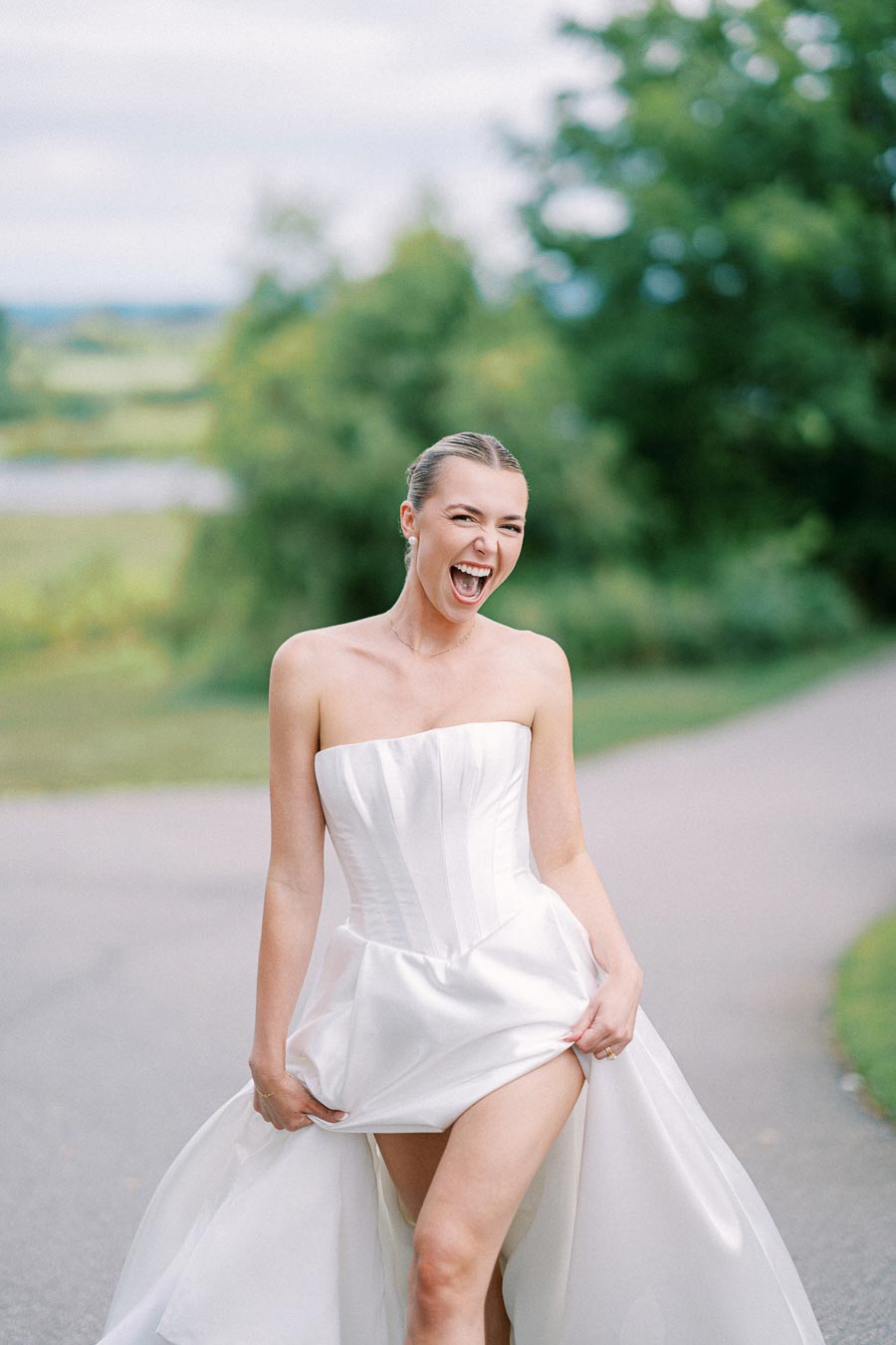 A joyful bride in a strapless white wedding gown laughing outdoors on a scenic pathway, surrounded by lush greenery.