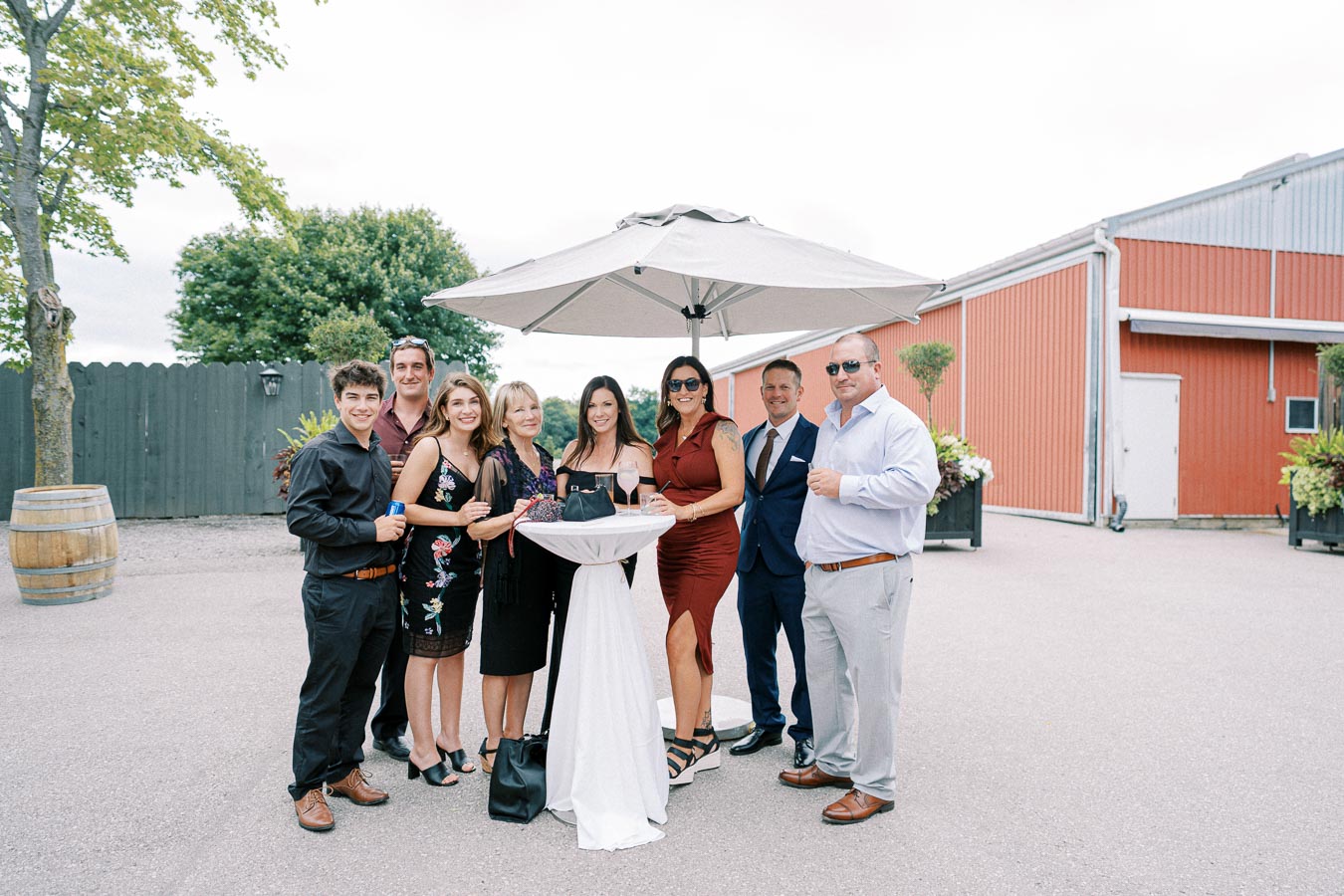 A group of people enjoying a social event outdoors under a large umbrella, standing around a cocktail table with drinks in hand. The setting includes a rustic barn and scenic greenery in the background.