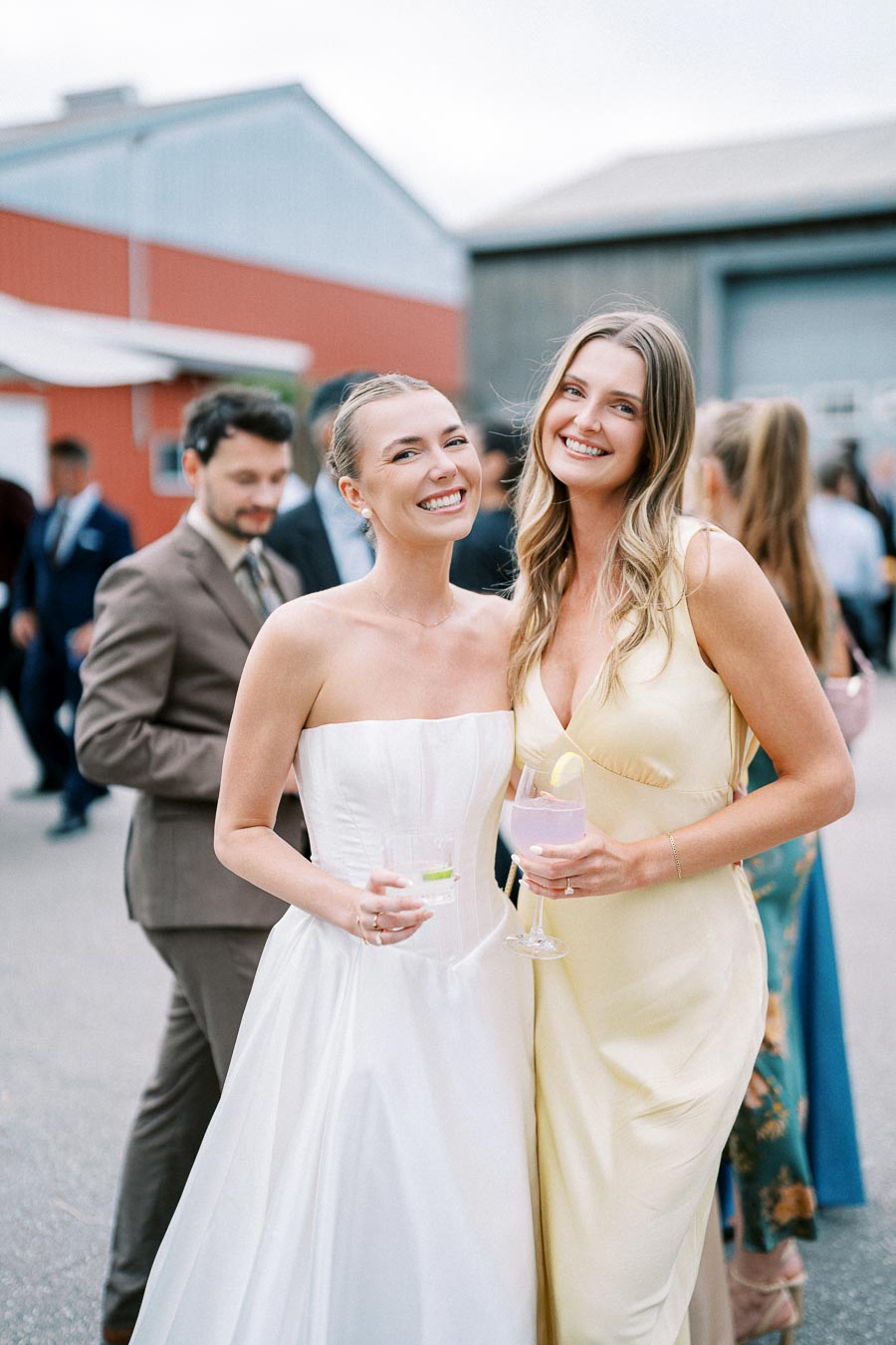Two smiling women in elegant dresses enjoying a wedding reception outdoors, holding drinks amidst a group of well-dressed guests.