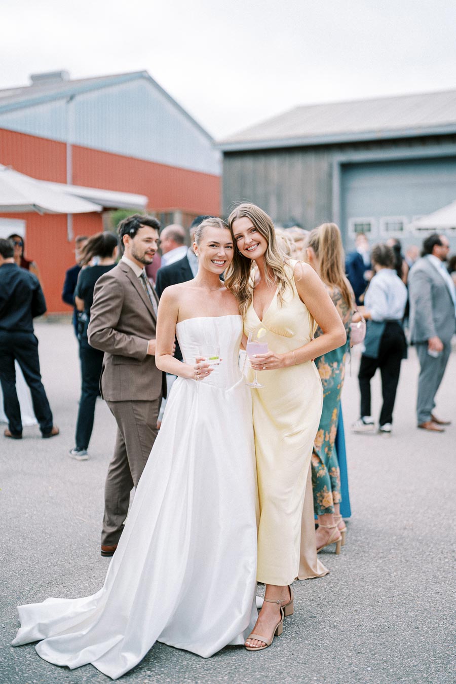 Two women smiling at an outdoor wedding reception, one in a white wedding dress and the other in a yellow dress, holding drinks with a crowd of guests and rustic building in the background.
