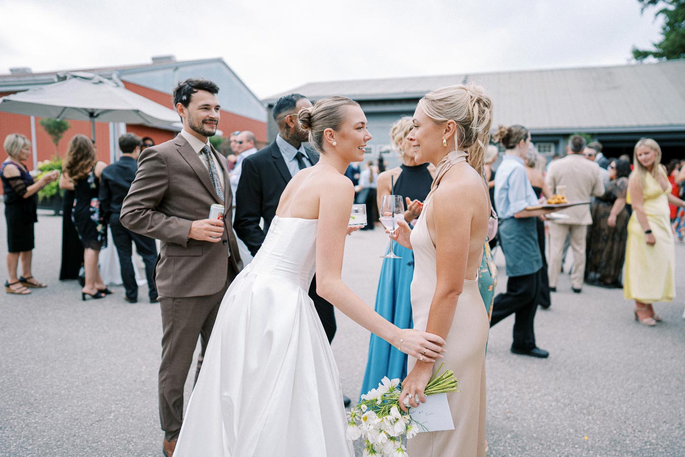 Outdoor wedding reception with guests socializing elegantly; a bride in a white gown converses with another woman holding flowers, surrounded by attendees in formal attire, creating a joyful and festive atmosphere.