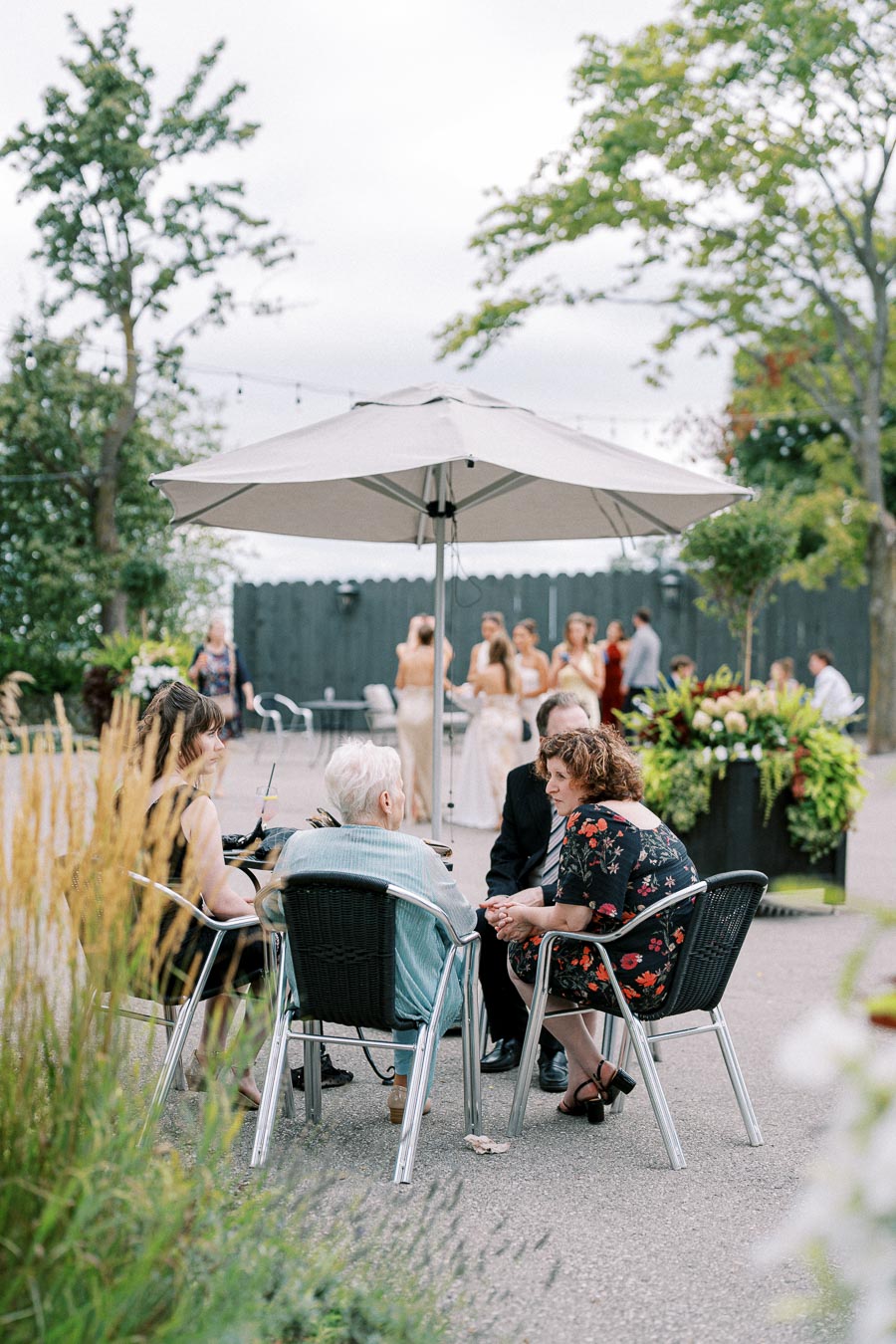 A group of people sitting at a table under a large outdoor umbrella, engaged in conversation at a garden event. In the background, elegantly dressed guests are gathered, with lush greenery and decorative planters enhancing the outdoor setting.