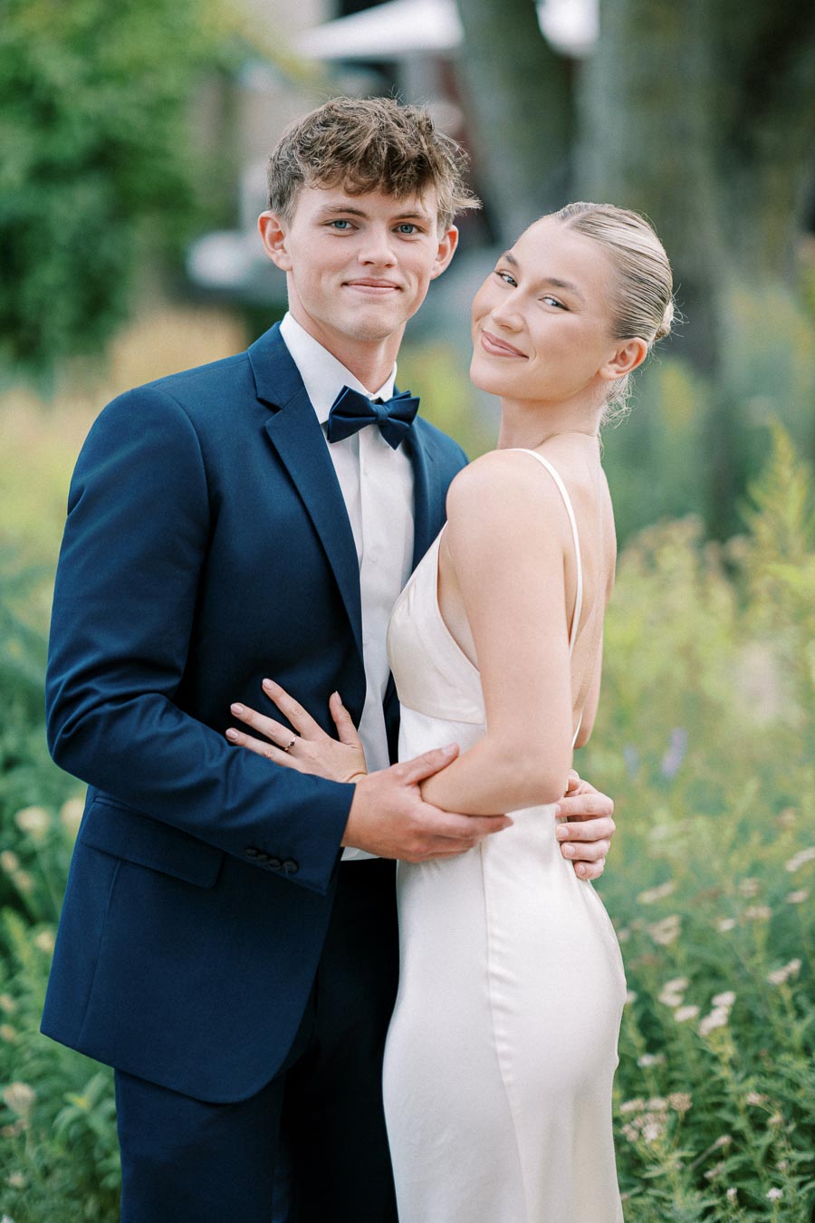 Smiling couple dressed in formal attire, posing in an outdoor garden setting, with the man in a dark blue suit and bow tie and the woman in a white sleeveless gown.