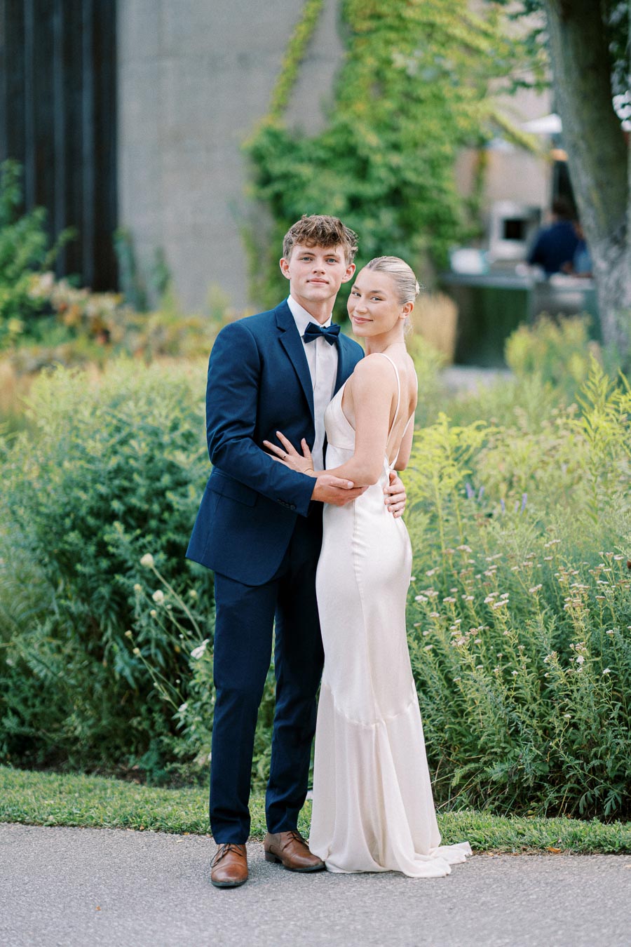 Young couple posing outdoors in formal attire, with the man in a navy suit and bow tie and the woman in a white evening gown, surrounded by lush greenery.