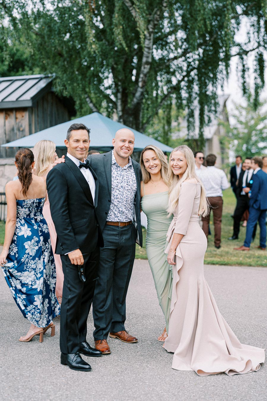 A group of well-dressed individuals smiling and posing outdoors at an elegant event, with men in suits and women in formal dresses, surrounded by lush greenery in the background.