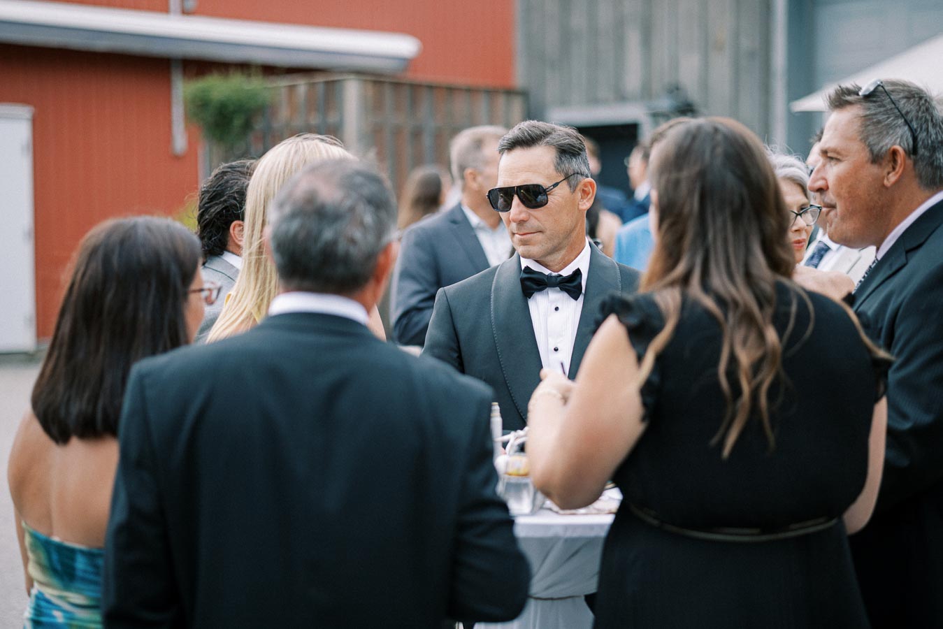 A group of elegantly dressed people socializing at an outdoor event, with a man in a tuxedo and sunglasses as the focal point, creating a sophisticated atmosphere.
