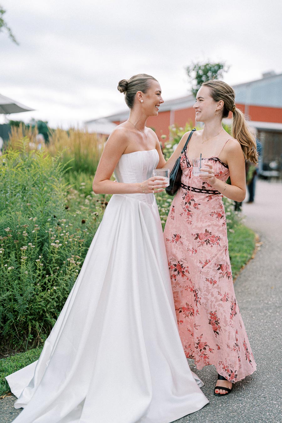 Two women at an outdoor event, one in a white wedding dress and the other in a pink floral dress, smiling and holding drinks, surrounded by lush greenery and a pathway.