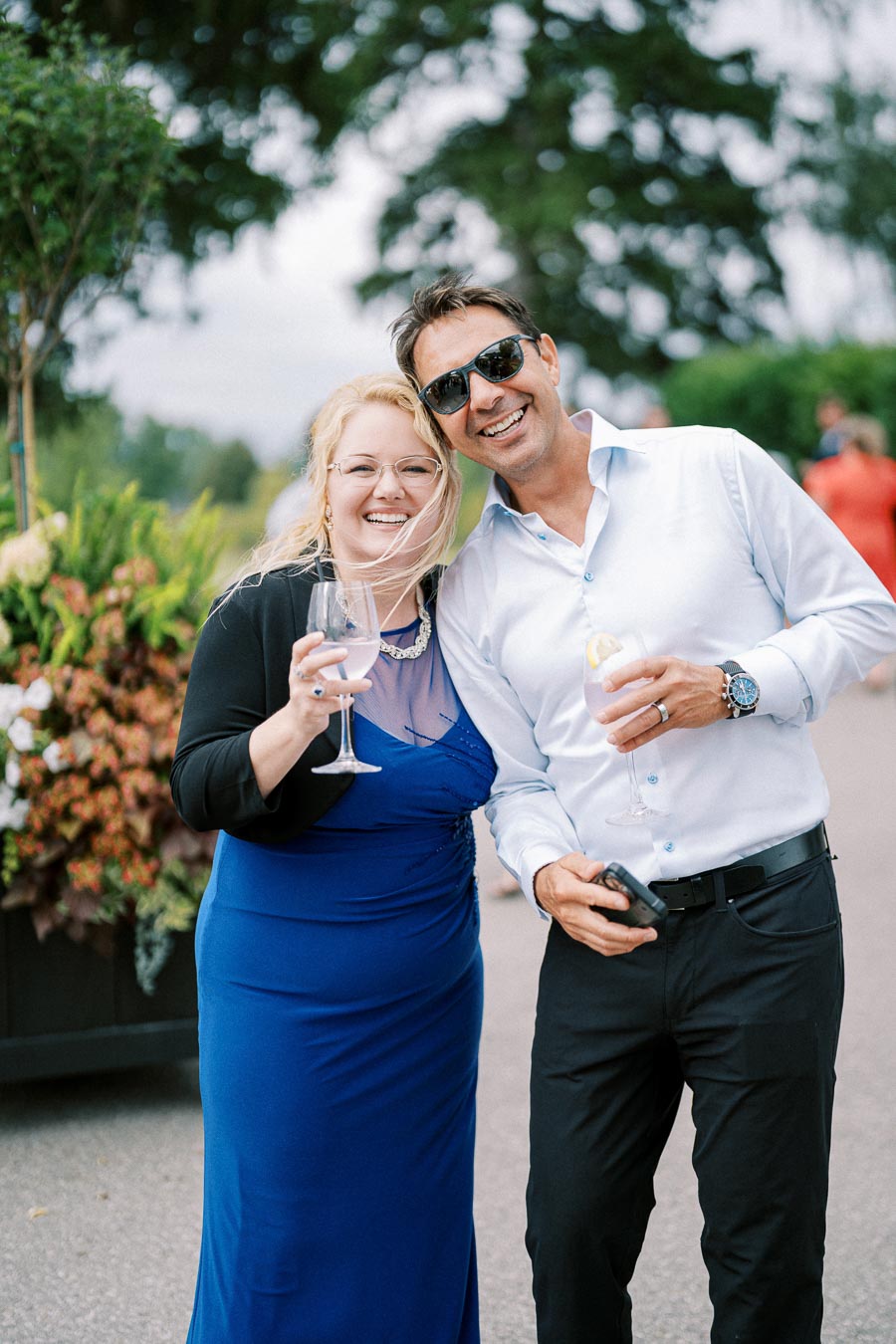 A smiling couple in formal attire enjoys a celebratory outdoor event, holding wine glasses, surrounded by lush greenery.
