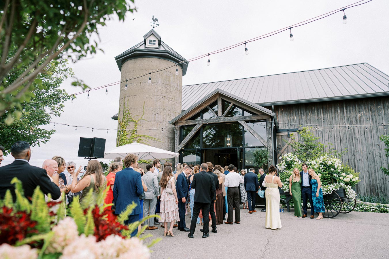 A lively wedding reception at a rustic barn venue, with elegantly dressed guests gathering outside, surrounded by vibrant greenery and flowers.