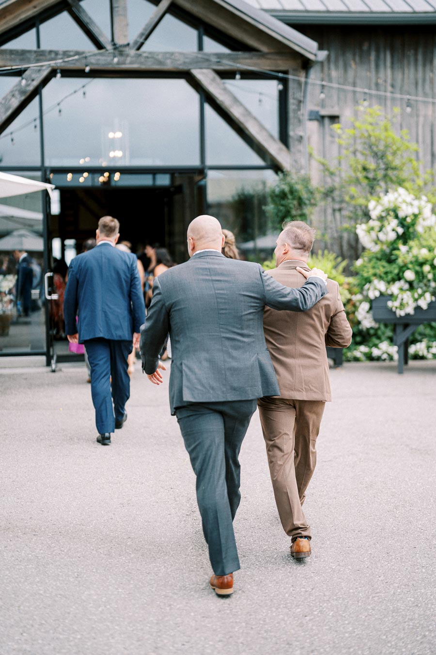Two men in suits walking towards a rustic wooden barn venue, with one arm around the other's shoulder, surrounded by greenery and soft lighting.