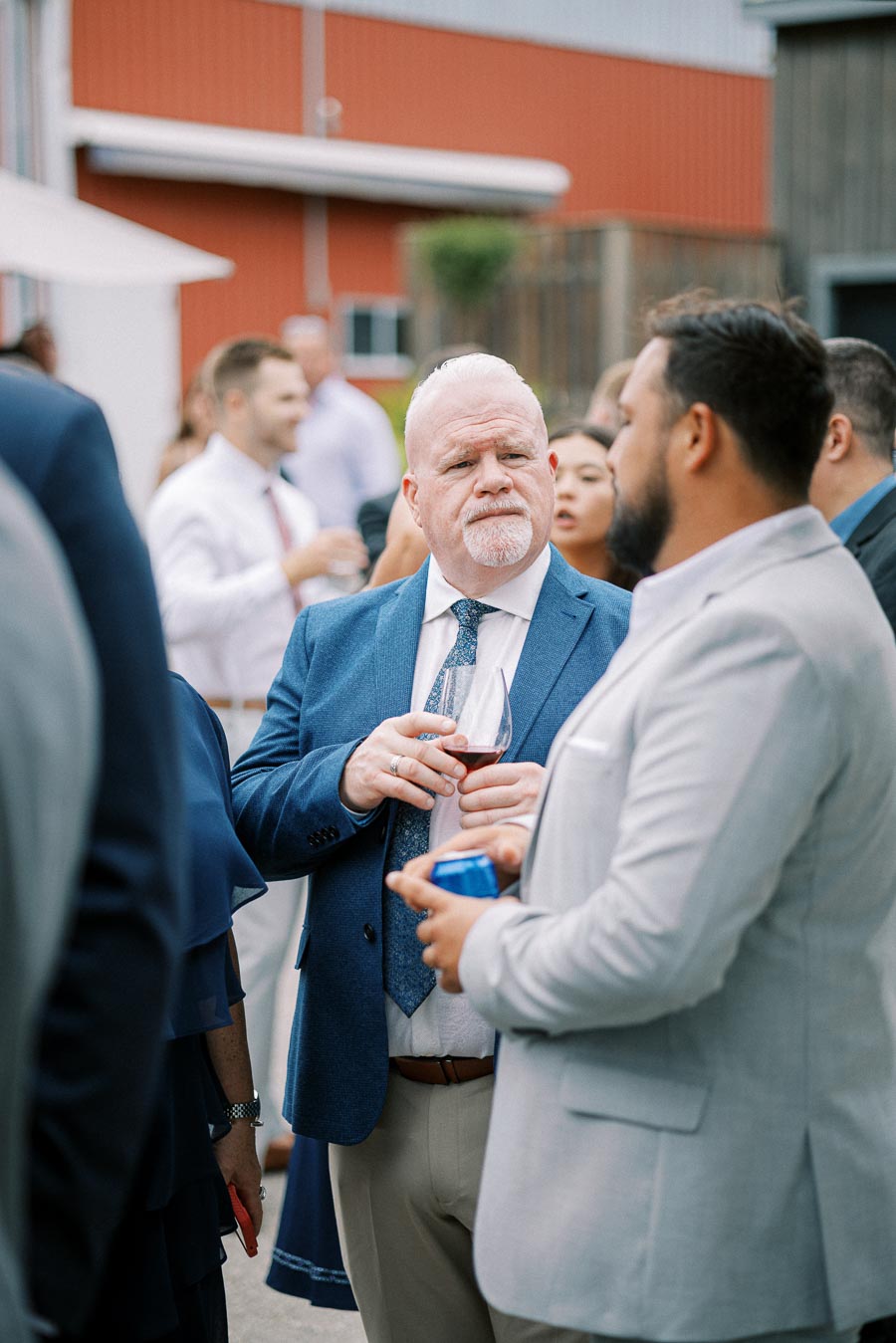 A man in a blue suit holds a glass of wine while conversing at an outdoor event.