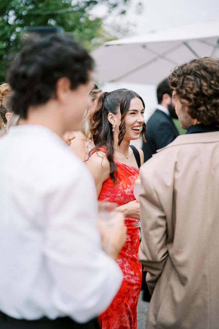 A woman in a vibrant red dress smiling and socializing at an outdoor event, surrounded by people in formal attire.