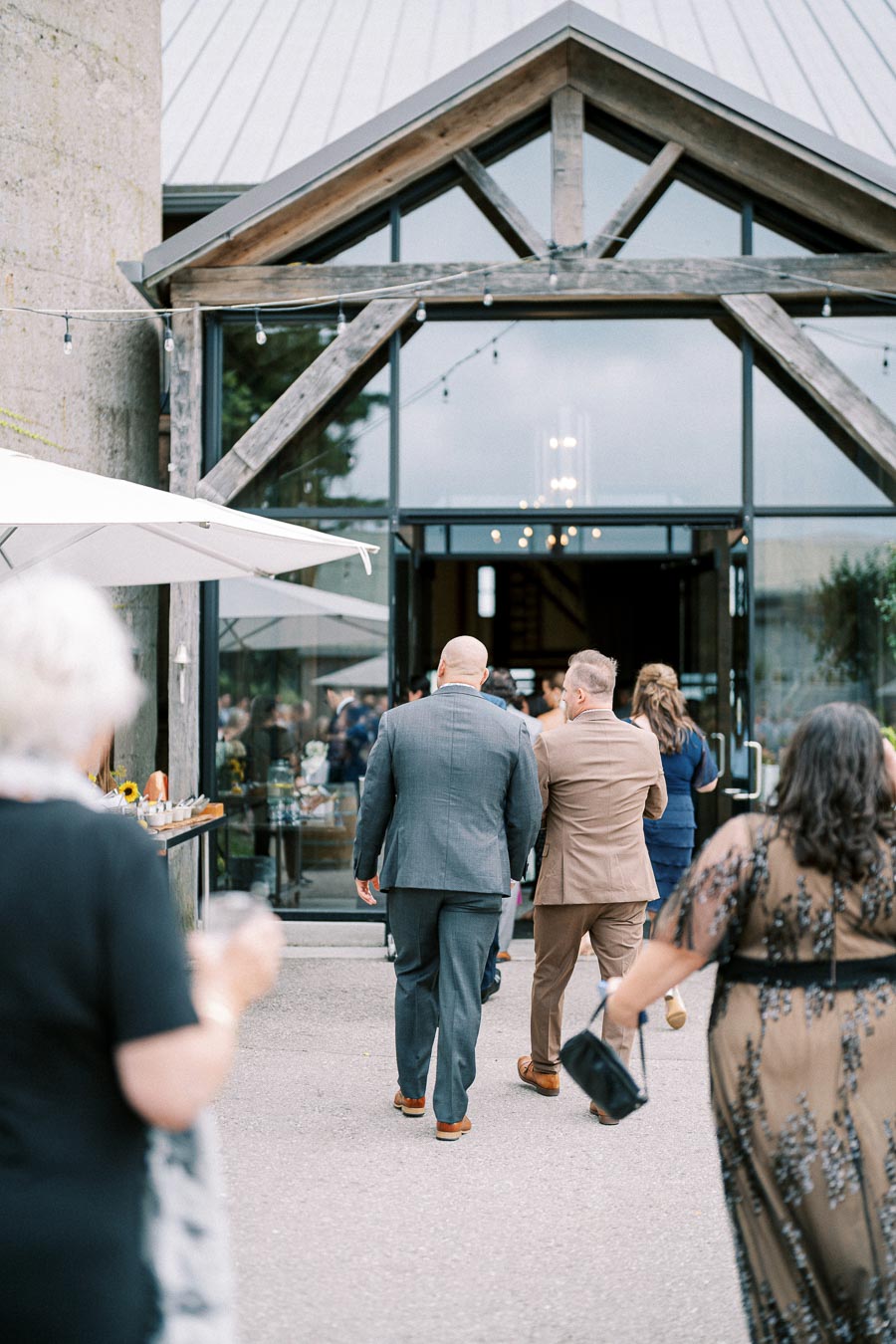 Guests entering a rustic venue with large glass doors at an outdoor event, featuring people in formal attire and decorative string lights.