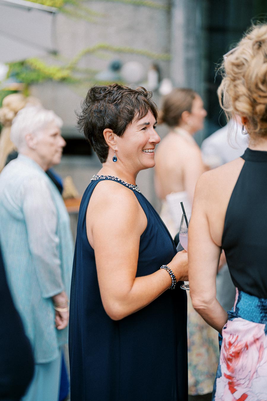 Elegant woman in a navy blue dress smiling and holding a cocktail at a social gathering, surrounded by other attendees, showcasing stylish event fashion.