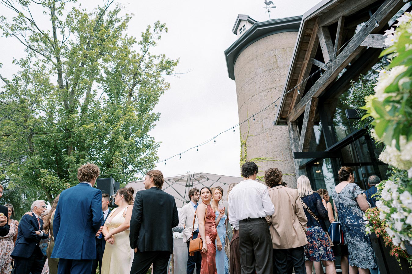Guests gathering at an outdoor wedding reception near a rustic building with string lights and lush greenery.