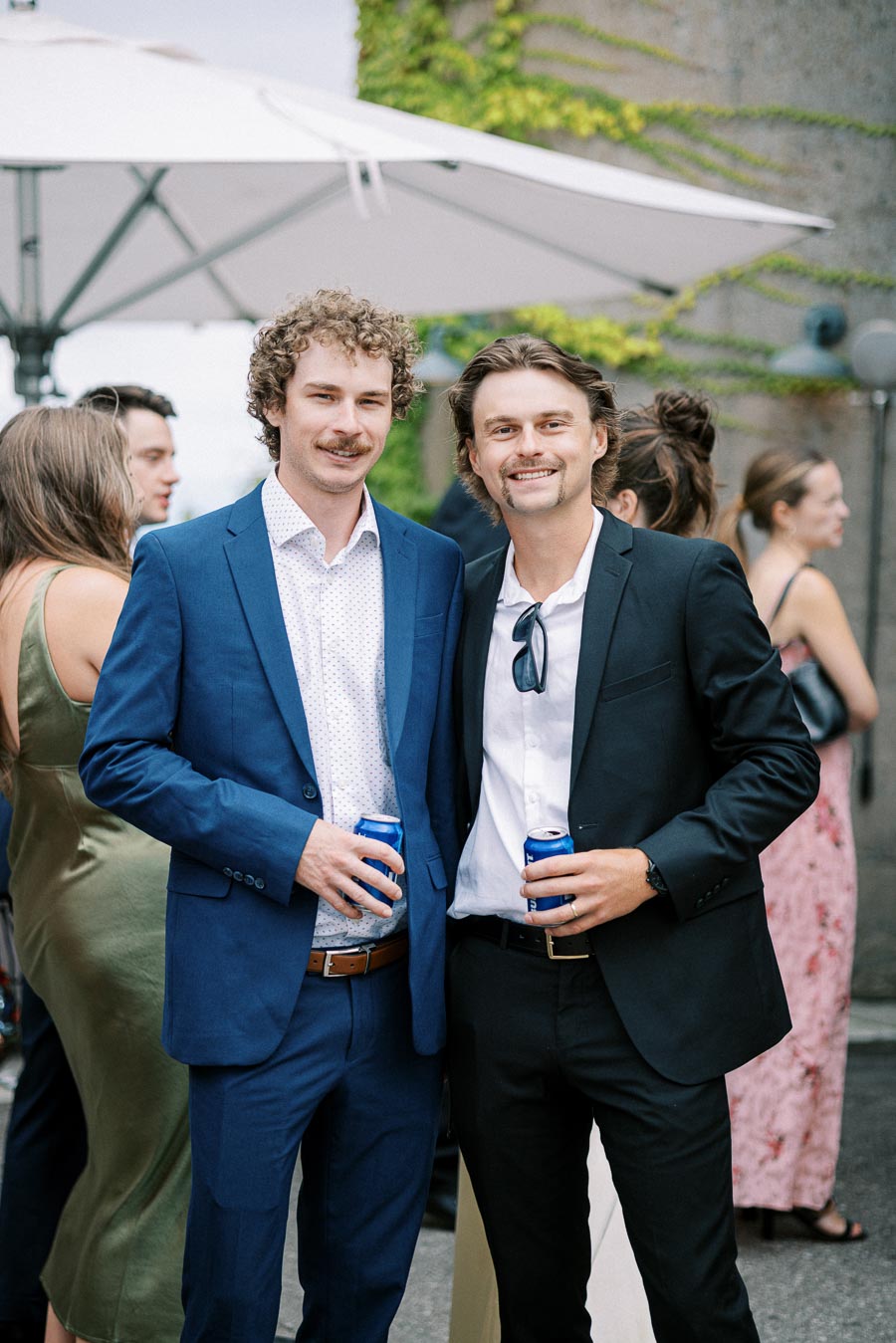 Two men in suits happily holding drinks at an outdoor social event, surrounded by other guests in casual attire.