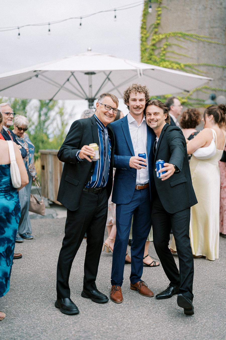 Three men smiling and posing with drinks at an outdoor social event, dressed in formal attire, with a background of people mingling under a large umbrella.