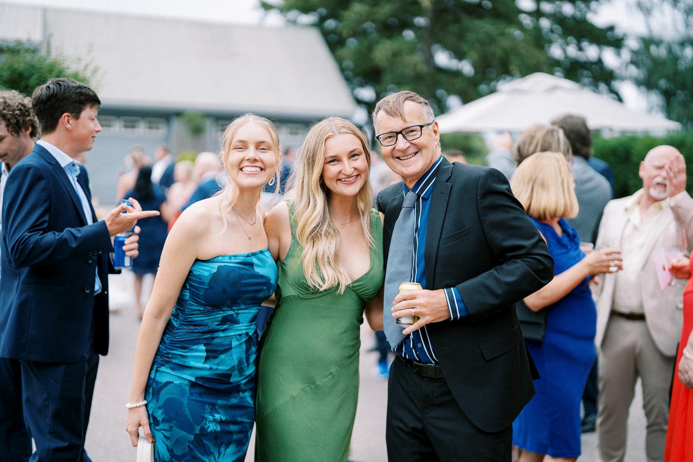 A group of people smiling at an outdoor event, with two women in elegant dresses and a man in a suit and tie, surrounded by a lively crowd enjoying the gathering.
