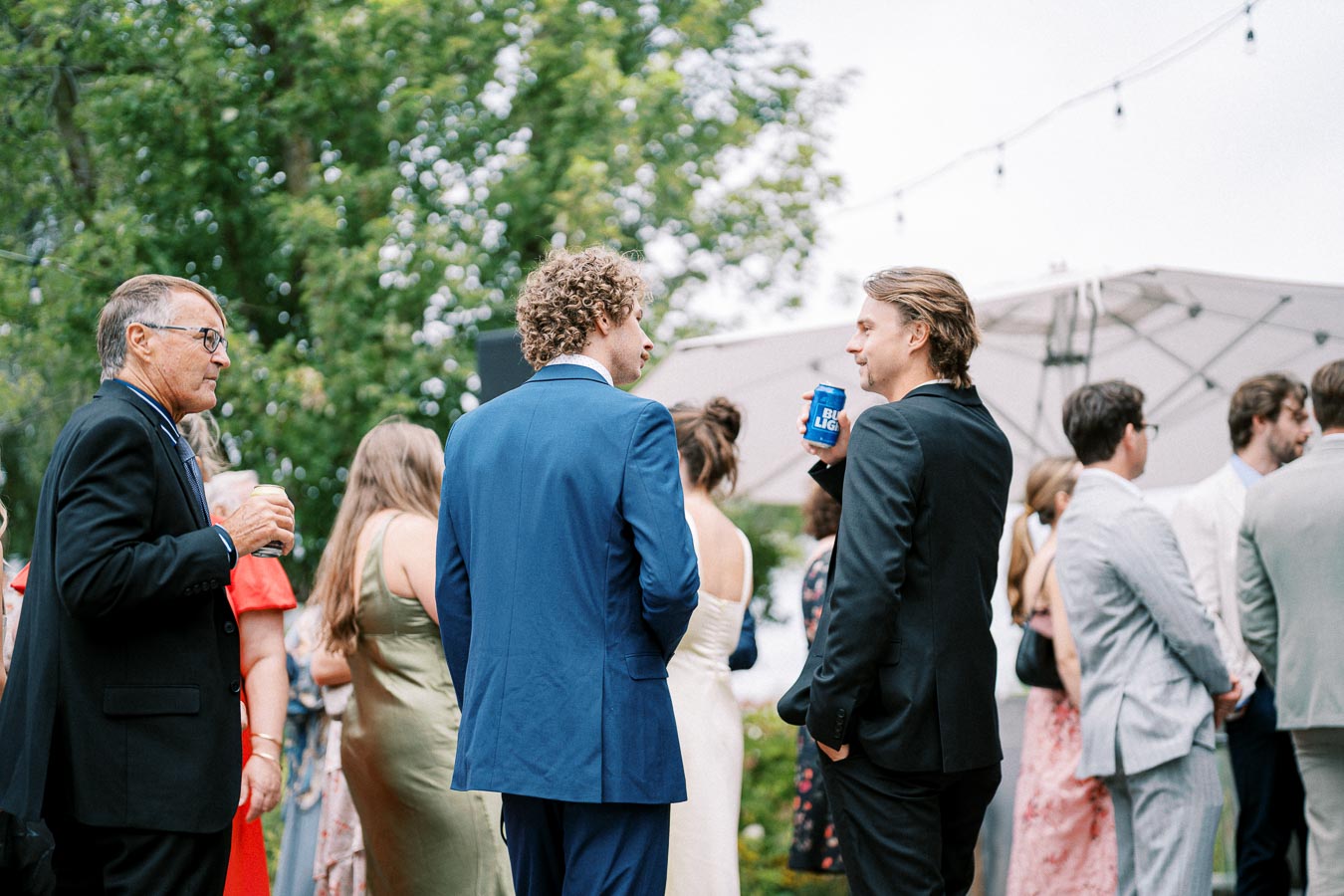 A group of people dressed in formal attire, socializing outdoors at a wedding reception, with string lights and greenery in the background. One man is holding a canned beverage.