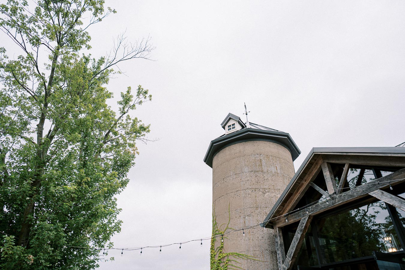 Rustic barn with a silage tower and lush green trees under an overcast sky, featuring string lights. Ideal setting for a countryside event or wedding venue.