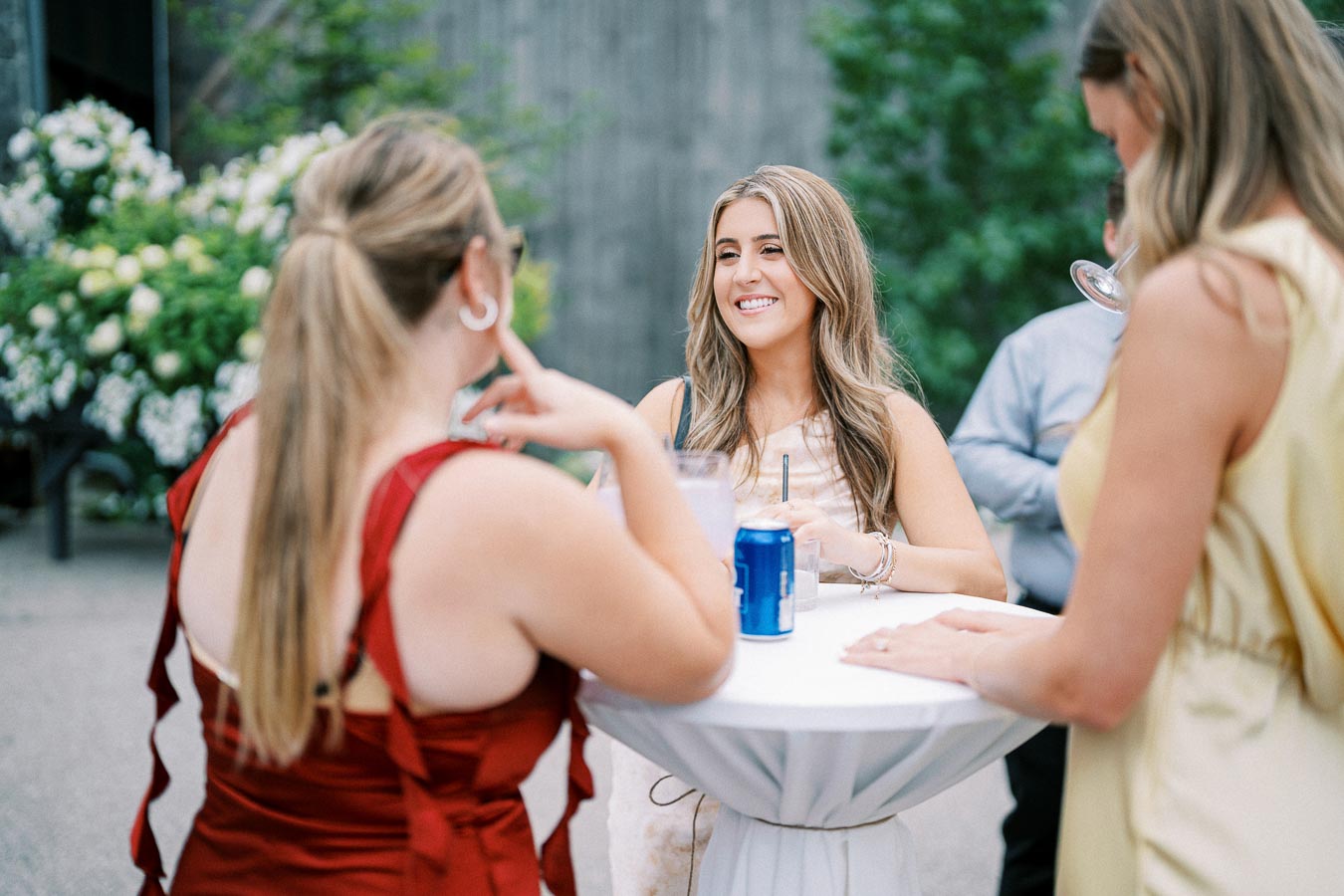 Outdoor social gathering with three women conversing around a cocktail table, drinks in hand, and greenery in the background