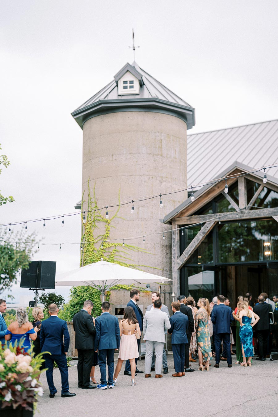 A lively wedding reception outside a rustic barn venue with guests in formal attire mingling under string lights, surrounded by greenery.