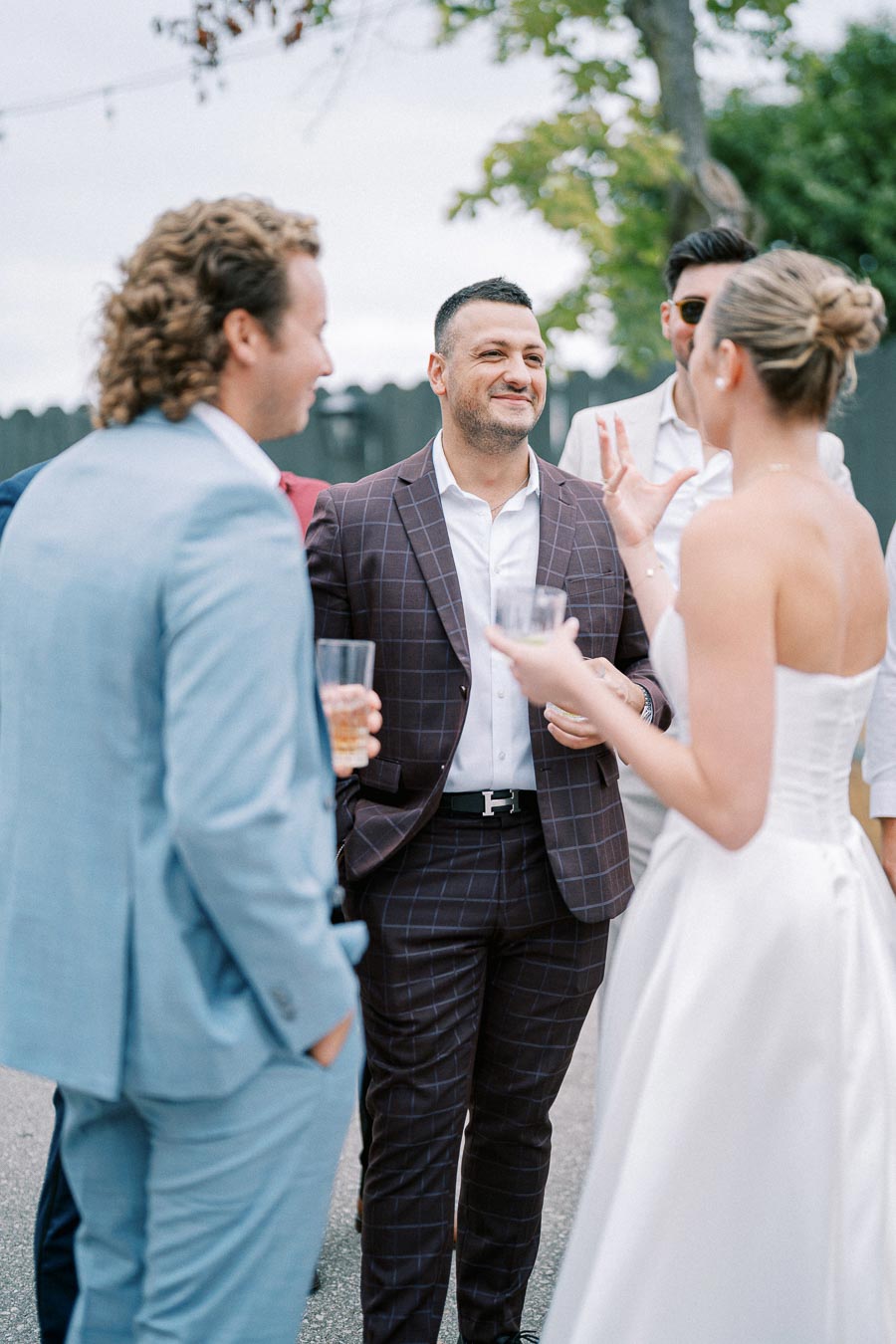 Group of well-dressed people socializing outdoors at a formal event, with a man in a checkered suit smiling and a woman in a white dress holding a drink.
