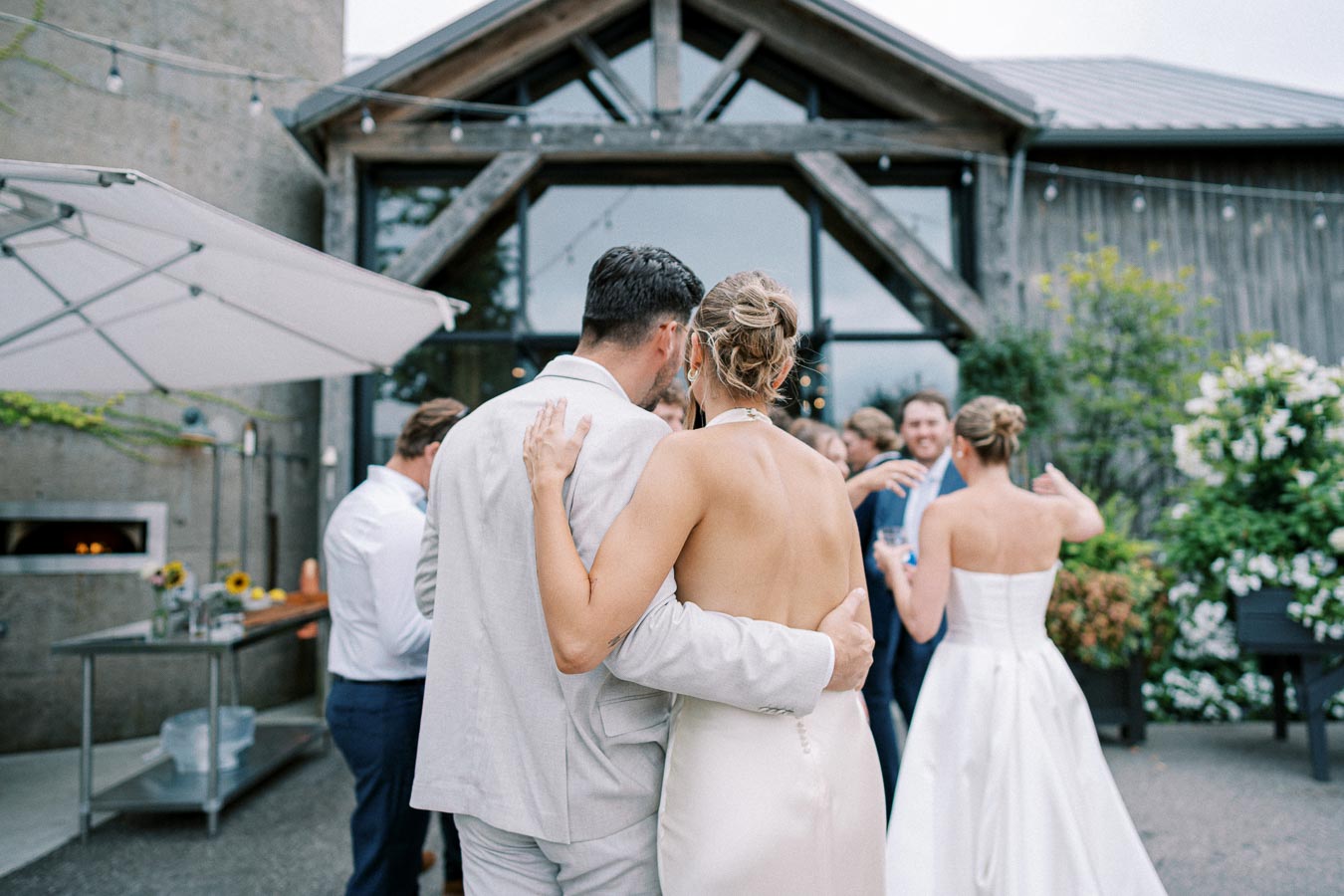 Outdoor wedding reception with a couple embracing in formal attire, surrounded by guests socializing in front of a rustic venue.