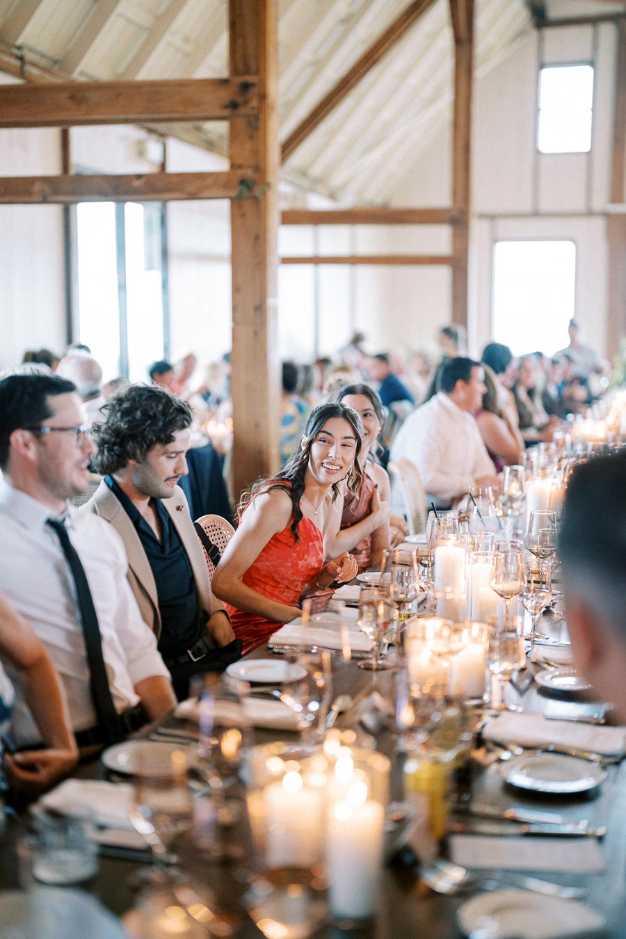 Guests enjoying a joyful wedding reception in a rustic venue, seated at a long, candlelit table with elegant table settings.