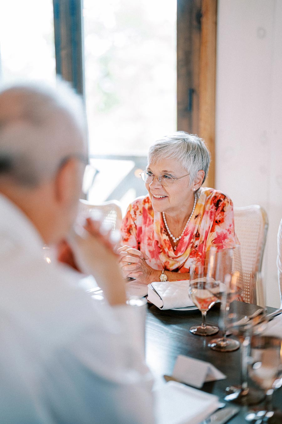 Senior woman with short gray hair and glasses, wearing a colorful floral blouse, engaged in conversation at a dining table with another person in focus. The table is set with glasses, cutlery, and napkins in a warmly lit room.