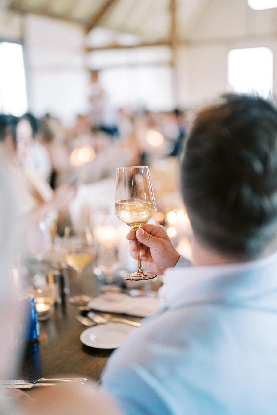A person raising a glass of white wine in a celebratory toast at a banquet with blurred guests and candlelit tables in the background.