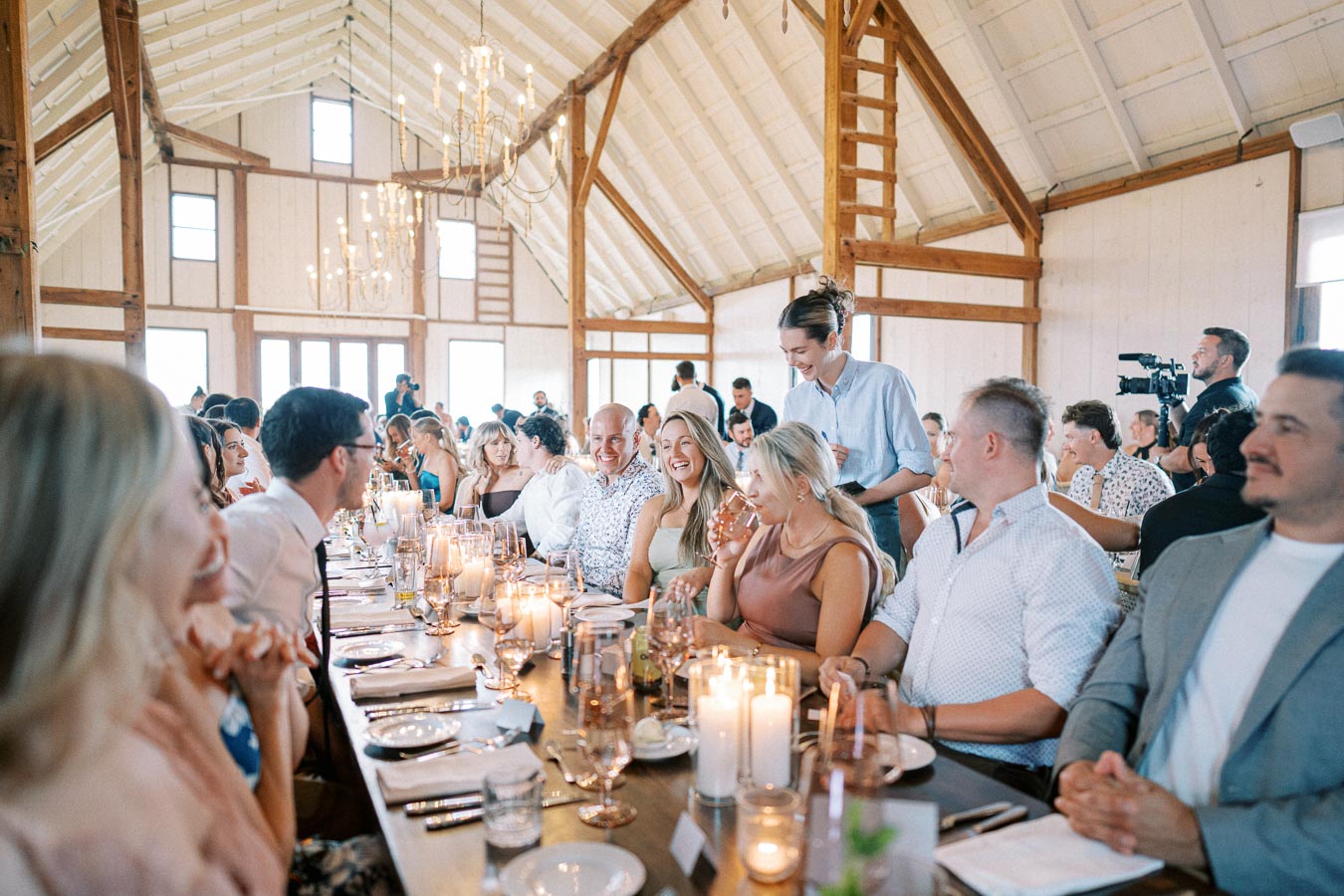Guests gathered around a long, elegantly set table in a rustic barn wedding venue, enjoying a meal with candles and laughter enhancing the warm ambiance.