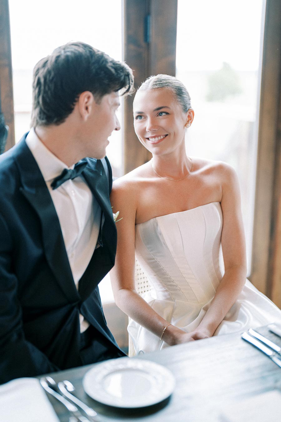 Elegant bride and groom smiling at each other during their wedding reception, bride in strapless white dress, groom in black tuxedo, seated at dining table.