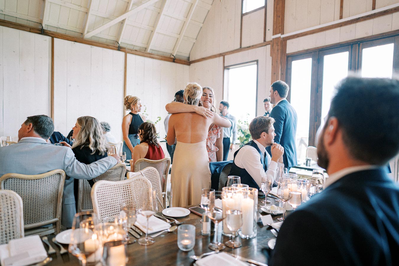 Elegant wedding reception scene in a rustic venue, showcasing guests happily interacting and celebrating. Tables are beautifully set with candles and fine dining ware, creating a warm and inviting atmosphere.