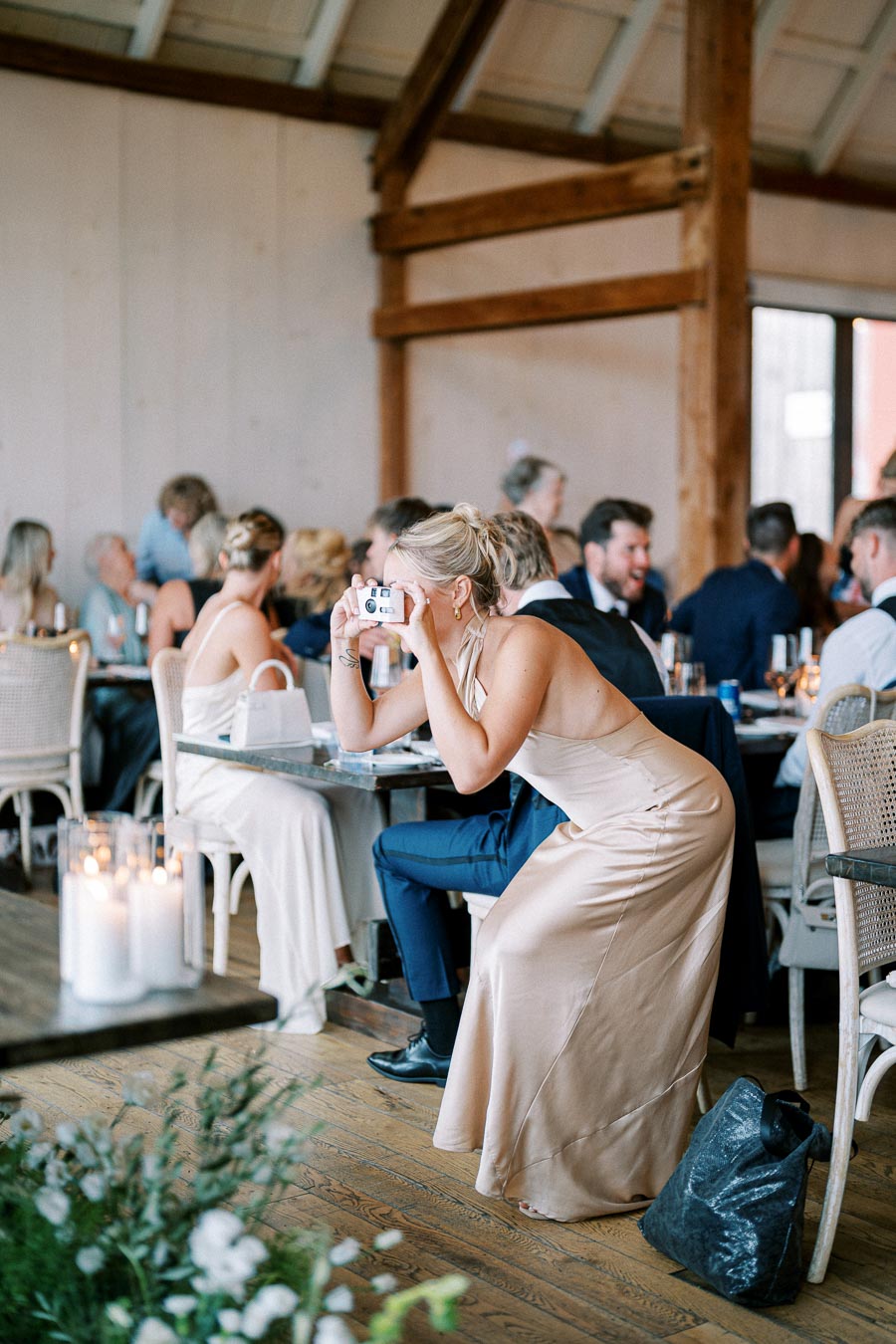 A woman in an elegant cream dress taking a photo with a disposable camera at a wedding reception, surrounded by guests seated at tables decorated with candles and flowers in a rustic venue.