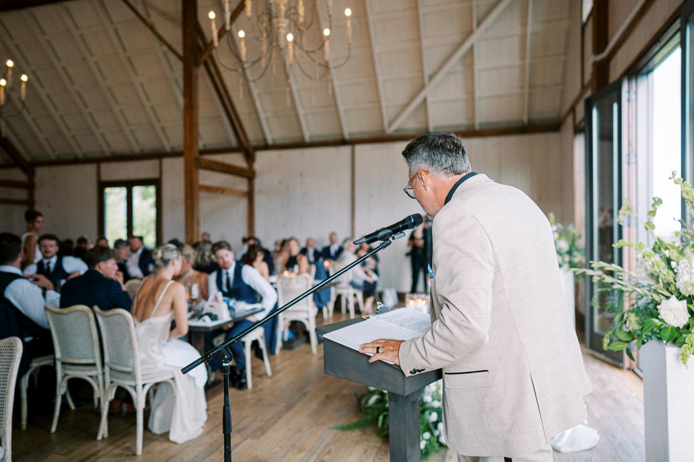A man in a beige suit giving a speech at a wedding reception in a rustic barn venue, with guests seated at tables in the background and floral decorations adorning the space.