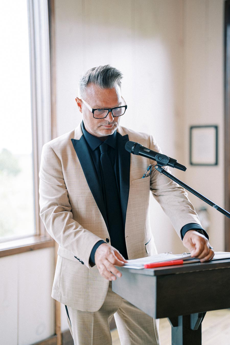 Man in a beige suit and black shirt speaking into a microphone at a lectern during a formal event indoors.
