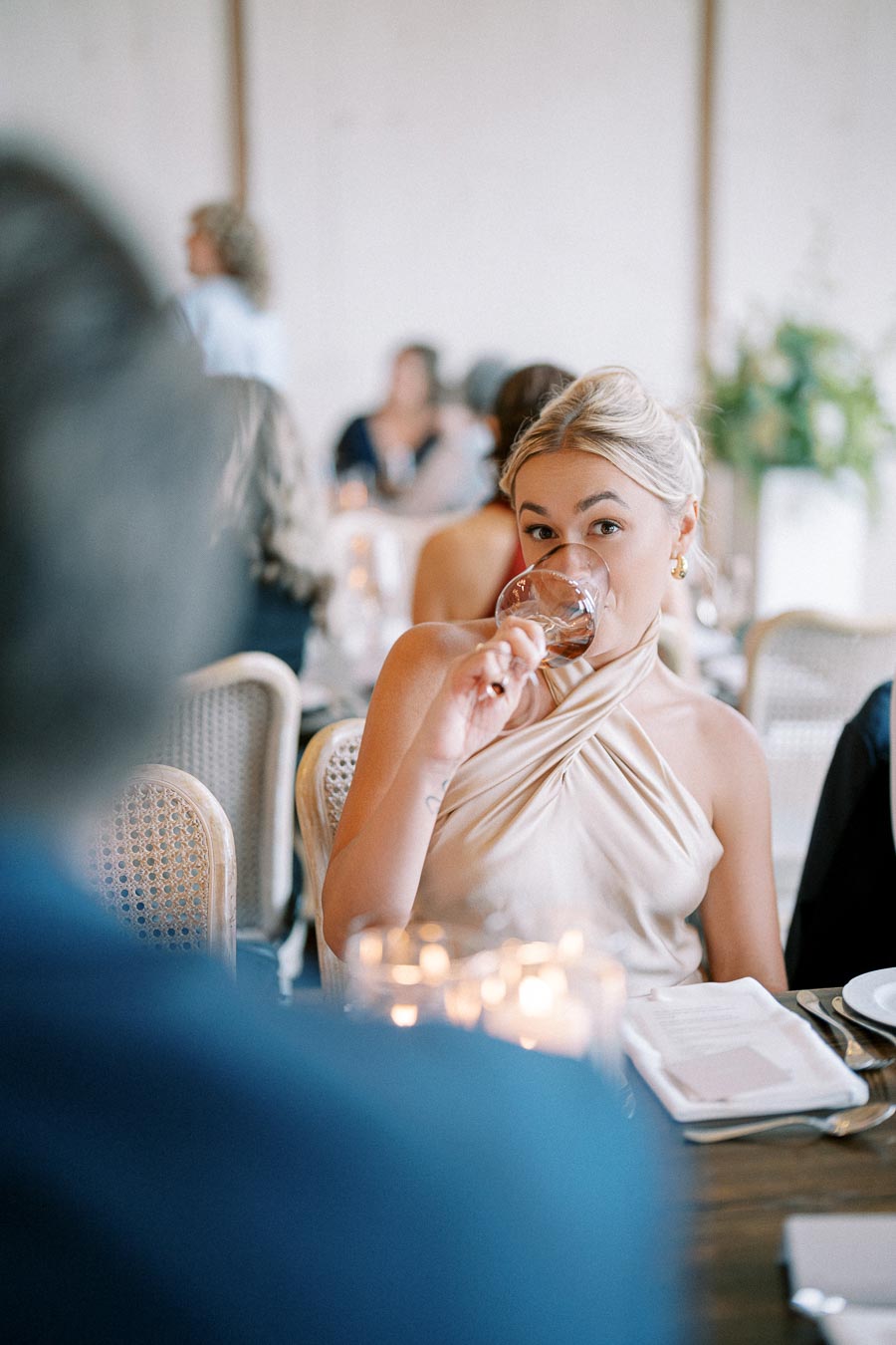 A woman with blonde hair sips a beverage from a glass at a formal event, dressed in an elegant halter neck dress, surrounded by softly lit candles and diners in the background.