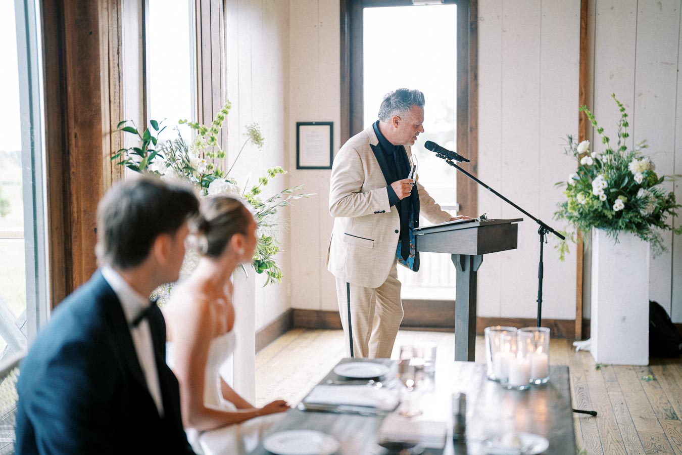 Wedding reception speech in a bright, elegant venue with speaker at a podium, floral arrangements, and a bride and groom seated in the foreground.
