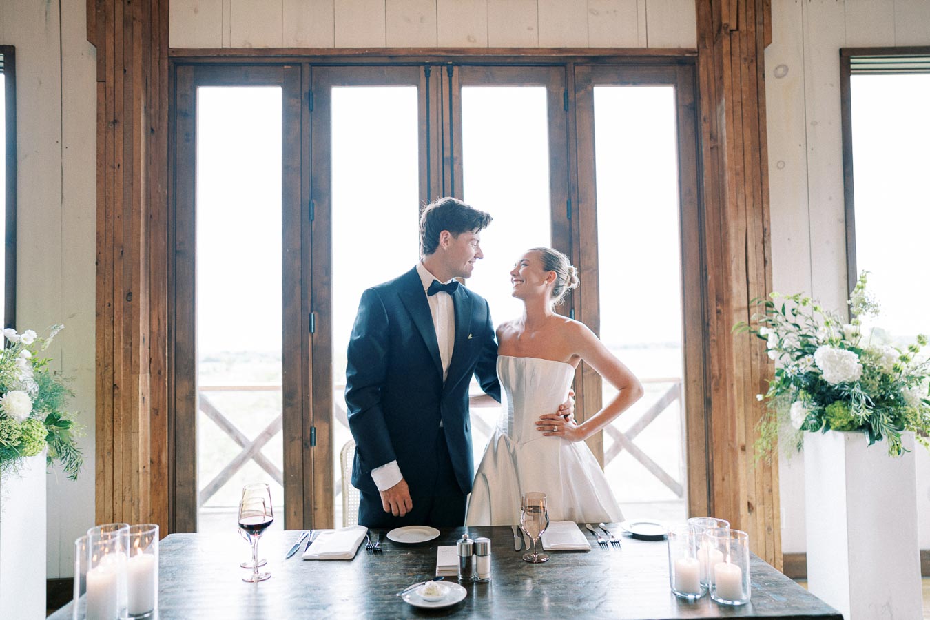 A bride and groom smiling at each other in a beautifully decorated wedding venue with candles and flowers.
