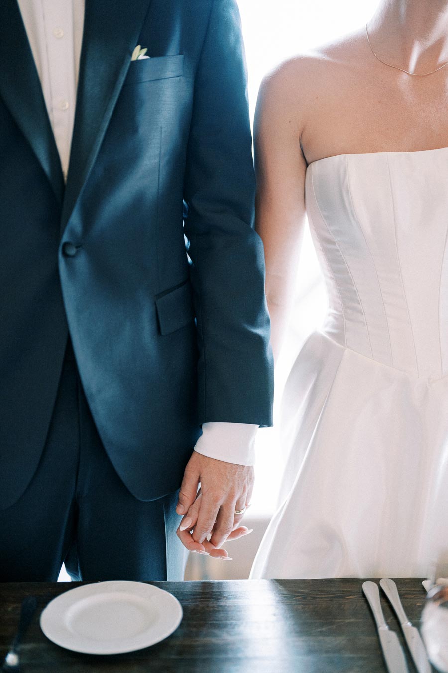 Elegant bride and groom holding hands at wedding reception table, dressed in formal attire, showcasing romance and commitment.