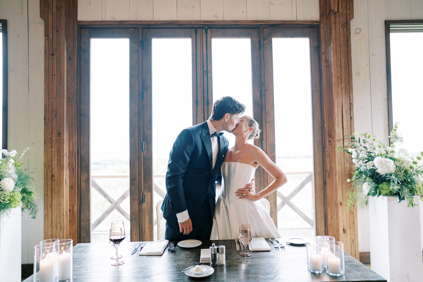 Elegant bride and groom sharing a kiss at a beautifully set dinner table with floral arrangements and candles in a rustic venue.