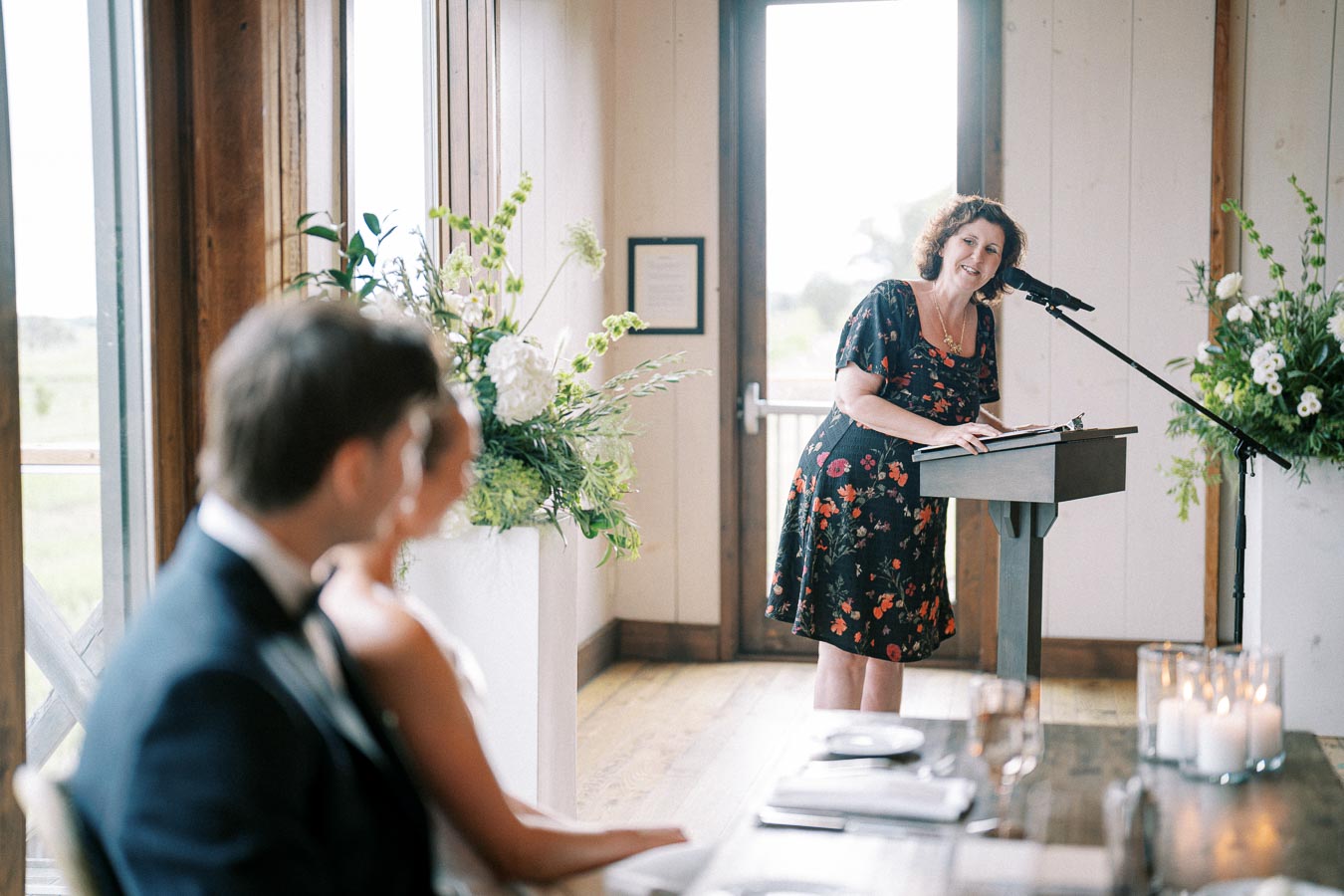 Wedding ceremony with a woman giving a speech at a podium, elegantly decorated with floral arrangements and candles, as a couple listens in the foreground.