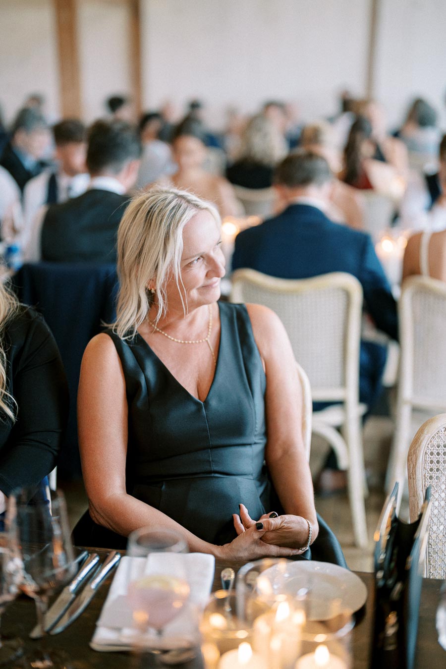 A smiling woman in a black dress sits at an elegantly set dining table, surrounded by blurred guests at a formal event or wedding reception.