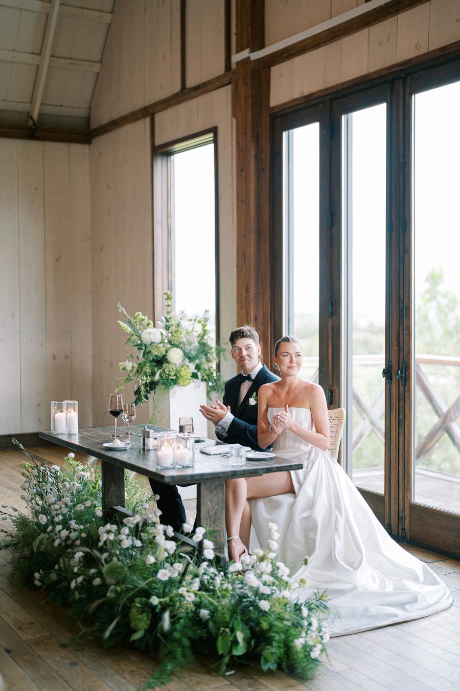 Elegant wedding reception with a bride and groom seated at a rustic wooden table, adorned with white floral arrangements and candles, in a light-filled venue with large windows.