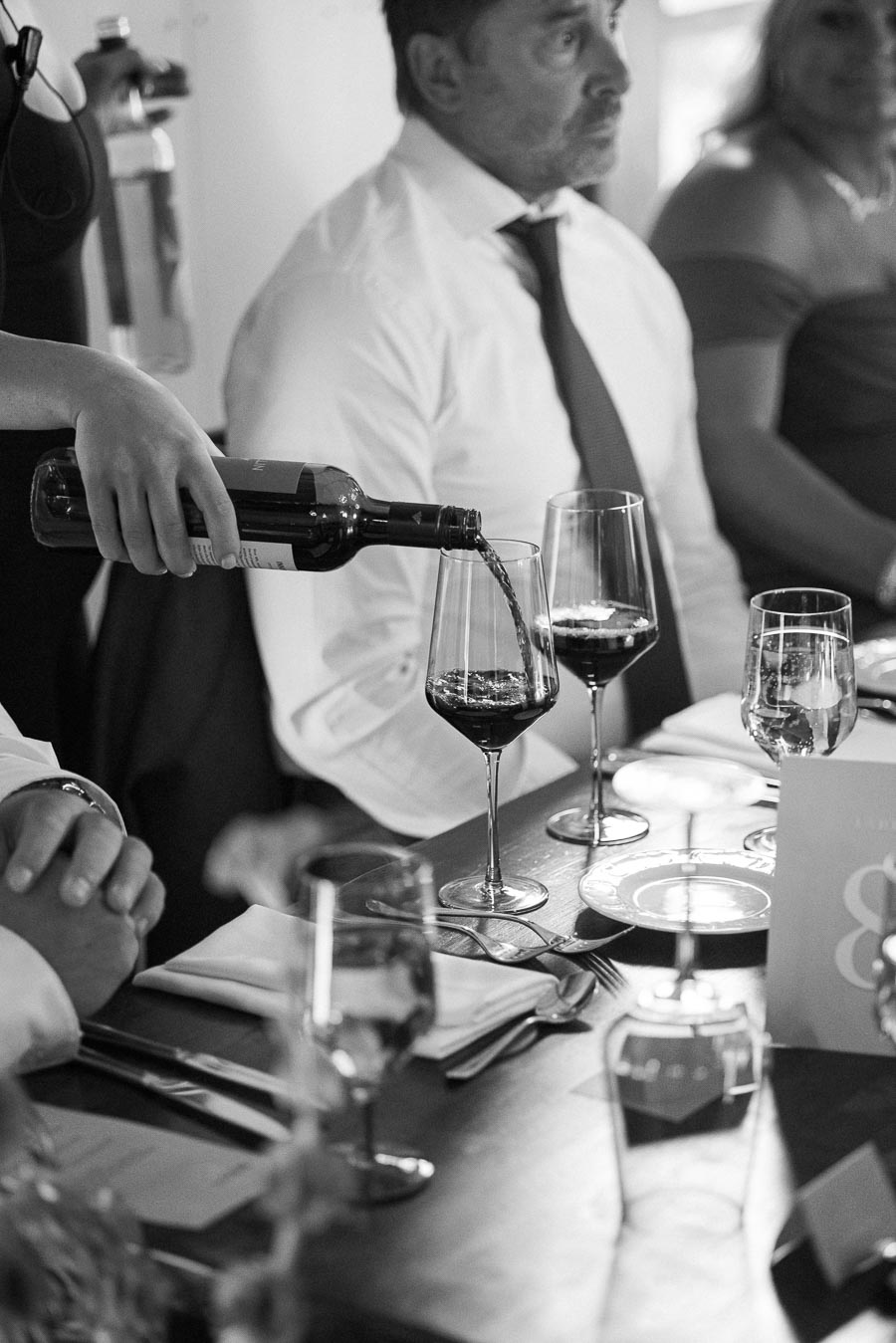 Black and white image of a formal dinner setting with a server pouring red wine into a glass, surrounded by attendees seated at a table set with elegant dining ware.
