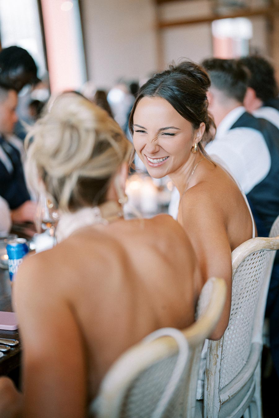 A woman smiling and laughing at a social event, dressed elegantly with her hair styled, surrounded by other guests in a warmly lit venue.